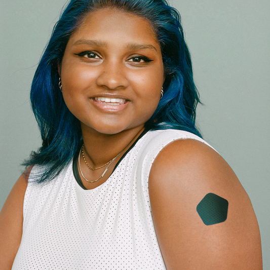 Smiling individual with blue hair wearing a sports top and a Recover patch on their arm for pain relief after needle procedures.