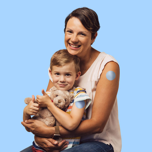 Mother and child smiling while holding a teddy bear, showcasing a comfortable Prepare patch on the child's arm.